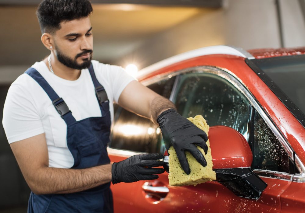 Young professional male worker in protective clothes and rubber gloves, holding a sponge for washing car rearview mirror and door. Selective focus on hand of man.